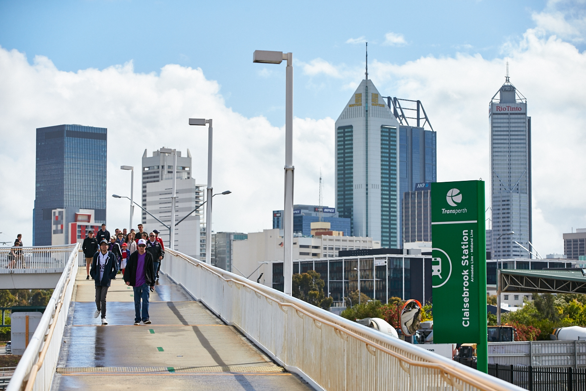 A photo of patrons walking down a pedestrian bridge with the Perth skyline in the background